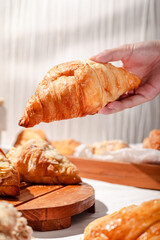 Fresh golden croissant held by a hand, showcasing its flaky texture and buttery layers, set against a rustic bakery background with almond pastries and wooden serving boards on a white tablecloth