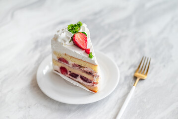 Slice of strawberry shortcake with layers of cream and fresh strawberries, garnished with whipped cream and parsley, served on a white plate with a gold fork on a marble table