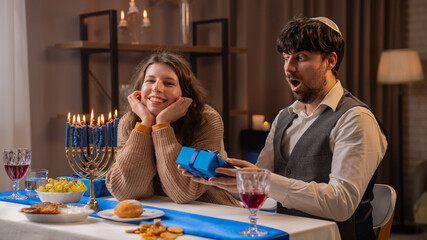 Jewish couple preparing for traditional hanukah celebrations, man and woman sitting at table, enjoying dinner and surprising with gifts, positive expression.