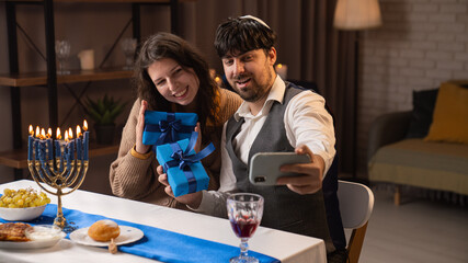 Jewish couple preparing for traditional hanukah celebrations, man and woman sitting at table, enjoying dinner, taking selfie with gifts on smartphone.