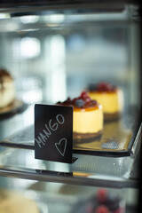 Row of portioned cheesecakes topped with mango cream cheese and fresh berries, displayed in a transparent dessert glass vertical display. A black charcoal tag with 'Mango' and a hand-drawn heart 
