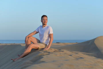 A young man in a desert at the sunset golden hour. A man wearing white t-shirt seating on a sand dune barchan at the background of the sea in Maspalomas, Gran Canaria