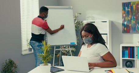 Man is standing at whiteboard holding tablet looking for right graphic preparing diagram beautiful woman is sitting at desk entering data into system needed to create diagram they are talking discuss