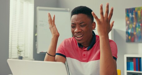 Young dark-skinned boy sits slumped in front of computer, props hands under face, bad news, losing...