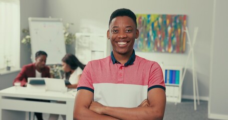 Obraz premium Portrait of man with dark eyes hair black skin beautiful wide white smile man is wearing salmon collar shirt student in classroom is looking at window then at camera coworkers talking in background