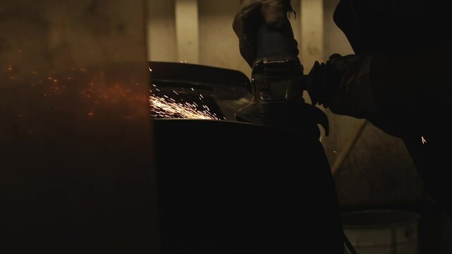 A man is using a grinding tool to work on a metal object. The image has a dark and moody atmosphere, with the man's silhouette and the shadows cast by the grinding tool. Concept of hard work