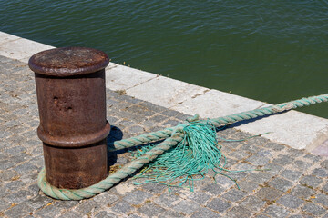 Fototapeta premium Rusty mooring bollard with green rope on cobblestones by the river