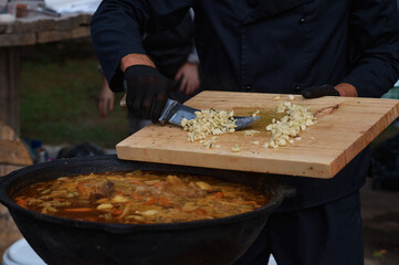 The chef adds garlic chopped on a cutting board to the meat soup with a knife. Boiling fresh food from vegetables and beef in a large cauldron. Catering and small business