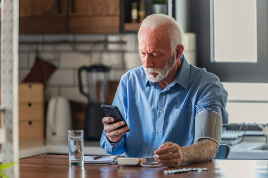 An elderly man with grey hair and a beard sits in his home kitchen, using a digital blood pressure monitor while checking his health data on a smartphone, looking focused and calm.