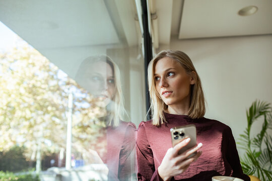 Young businesswoman holding smartphone looking through office window