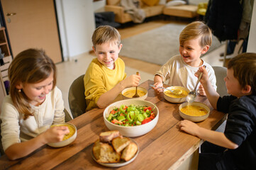 Children enjoy a hearty meal at the table, eating pea soup with croutons and fresh salad. They savor their food with appetite, creating a warm and cozy family dining atmosphere.