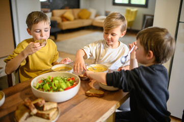 Children enjoy a hearty meal at the table, eating pea soup with croutons and fresh salad. They...