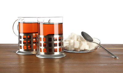 Glasses of aromatic tea in holders, sugar cubes and spoon on wooden table against white background