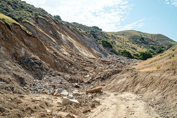 A rugged landscape featuring a large landslide on a hillside, scattered with rocks and debris, illustrating erosion and the impact of natural geological processes under a bright sky.