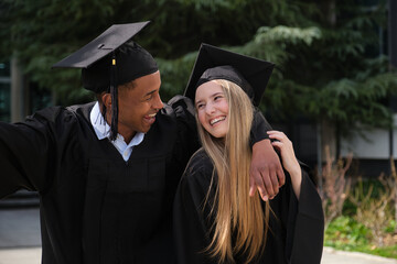 Two joyful multi ethnic students wearing graduation gowns are celebrating their academic achievement