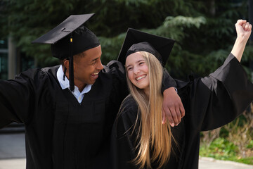 Two cheerful graduates wearing their gowns are celebrating their academic achievement outdoors