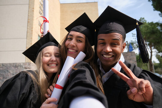 Group of cheerful multi ethnic graduated students taking selfie and showing peace sign