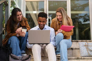 Multi ethnic university students using laptop and laughing together sitting on steps