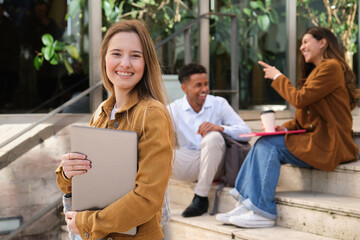 Portrait of a smiling female student holding a laptop standing in front of the university building while her classmates are talking and having fun sitting on the stairs