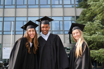 Three happy multi ethnic graduates wearing their gowns and mortarboards are smiling in front of...