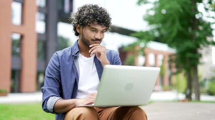 Young thoughtful businessman using laptop sitting on a bench near office building. Freelancer thinks about solution a problem working on computer. Student got an idea, found the answer. Close up