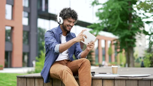 Happy male student in wireless headphones plays a video game on a tablet while sitting on bench in campus space near university building. Excited young handsome male enjoys an interesting online game