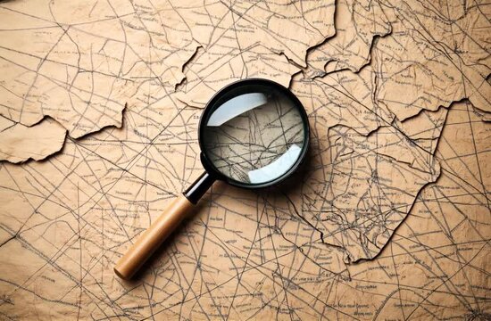 Vintage-style map with black lines and text on brown paper, partially torn. A magnifying glass with a wooden handle lies on top, magnifying part of the map.