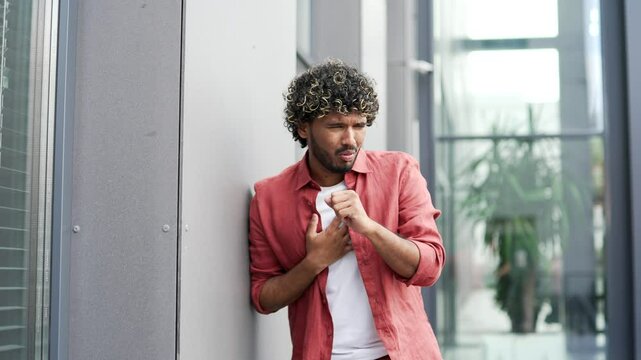 Sick young businessman coughing while standing on street near office building. Upset unhealthy handsome male with a cold or flu feels bad, holds his hand to his chest. He has bronchitis or pneumonia