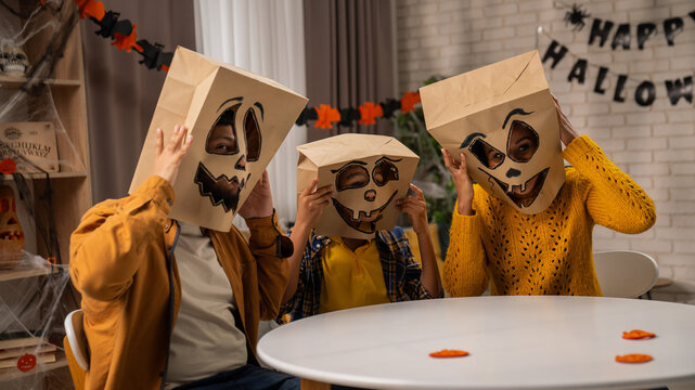 African american family of mom, dad and little son preparing for halloween celebrations, sitting at table taking selfie on smartphone in scary paper bag masks.