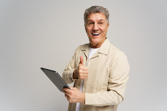 Older man smiles and gives a thumbs up while holding a tablet in a neutral backdrop