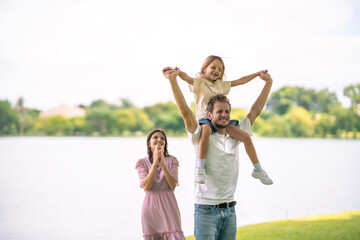 Cute White girl sitting on her father shoulder at park. Dad carry his daugther on his neck walking at pubplic park, mother crab hand to encourage them.
