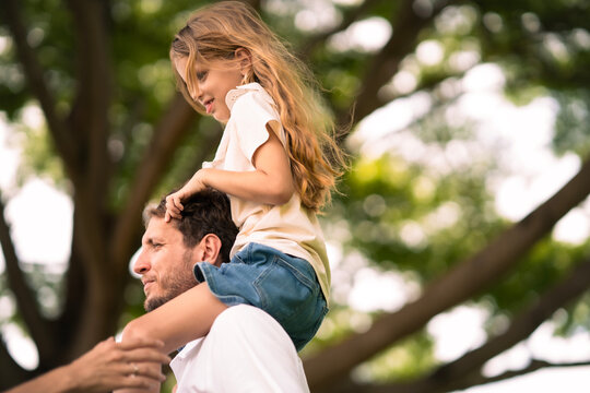 Cute White girl sitting on her father shoulder at park. Dad carry his daugther on his neck walking at pubplic park.
