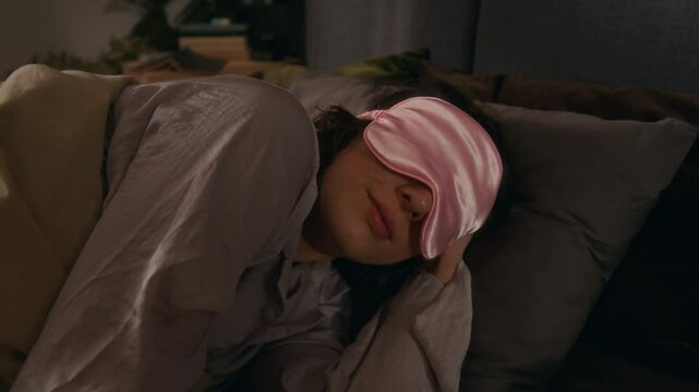 Young woman in pink sleep mask tossing, while sleeping in comfortable ned in modern eco-friendly bedroom
