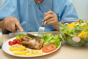 Asian senior woman patient eating pork chop stake and vegetable salad for healthy food in hospital.