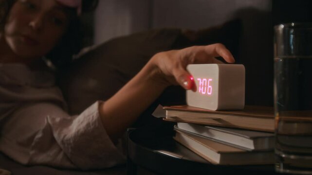 Woman taking off her pink sleep mask and picking up digital alarm clock from cluttered bedside table to turn it off, while lying in bed