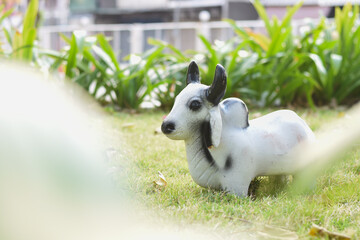 A white cow statue with black horns sits on a lawn with trees and green plants in the background.