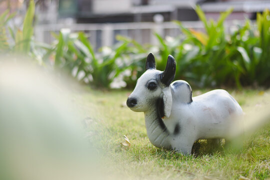 A white cow statue with black horns sits on a lawn with trees and green plants in the background. - Powered by Adobe
