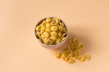 Top view of a porcelain bowl of raw italian durum wheat orecchiette pasta against a light ochre...