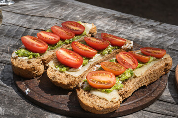 avocado and tomato bruschetta on whole grain bread, served on a wooden plate outdoors. The dish features creamy avocado spread, melted cheese, and vibrant cherry tomatoes.