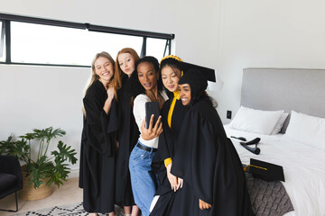Diverse graduates wearing caps and gowns taking selfie together in bedroom, celebrating, at home