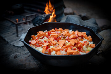 A black cast iron pot filled with fresh chopped vegetables sits atop a weathered wooden surface in a natural outdoor setting. The warm glow of the ingredients contrasts with the cool forest background
