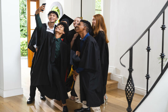 Diverse graduates in caps and gowns taking selfie, celebrating together at home