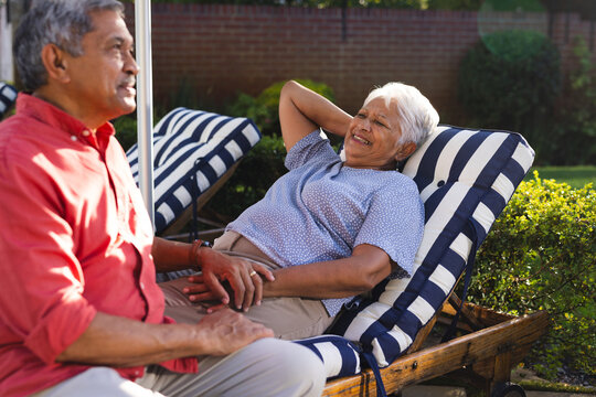 Senior diverse couple relaxing on patio chairs, enjoying conversation in sunny backyard