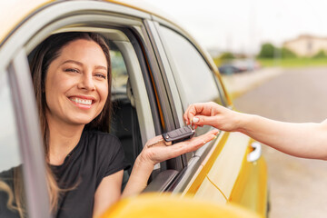 A smiling woman sitting in a yellow car receives a set of keys from another person, symbolizing a car purchase, rental, or ownership transfer.