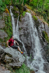 A man is peacefully sitting on a large rock near a beautiful waterfall