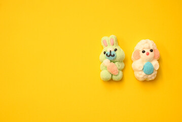 Easter-themed marshmallow figures—a fluffy lamb with an egg and a smiling green bunny with a carrot—on a vibrant yellow background. Perfect for seasonal designs.