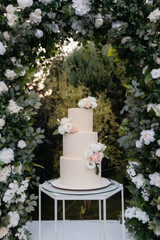 Trendy expensive wedding cake on a white arch of fresh flowers and sunset for a wedding ceremony