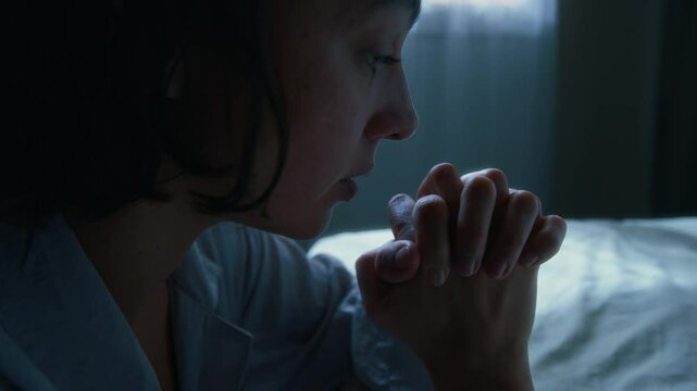 Close-up shot of young woman with dark short hair kneeling before bed at night and saying prayers before going to sleep