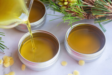 Pouring melted homemade pine needle salve balm into reusable tin container on white background.