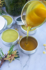 Pouring melted homemade pine needle salve balm into reusable tin container on white background.
